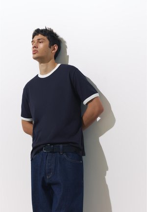 Mladić u tamnoplavim trapericama i mornarsko plavoj majici s bijelim rubom naslonjen na bijeli zid, gleda lijevo.