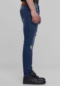 Tummansiniset skinny-farkut, joissa on kulutettu yksityiskohta polvissa, yhdistettynä mustiin korkeavartisiin tennareihin. Vyötäröllä on nahkapatche.