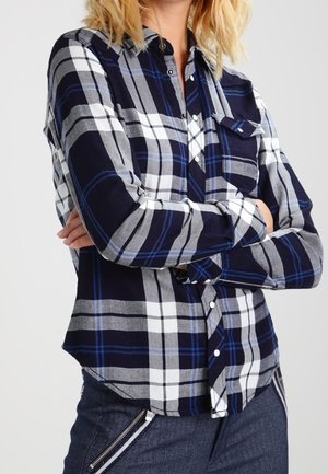 Persoon die een marineblauw, wit en grijs geruit overhemd met knopen draagt, met de armen gekruist, gecombineerd met een donkere broek met een ritssluiting detail.