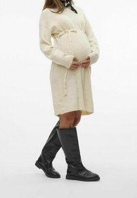 Robe tricotée de couleur crème avec une ceinture à nouer, portée avec de hautes bottes en cuir noir. La robe présente des manches longues et une coupe décontractée.