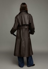 Brązowy skórzany trencz z paskiem w talii, długimi rękawami i klasycznym krojem. Posiada wysoki kołnierz i sięga poniżej kolan.