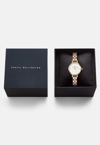 Zeltīts un sudrabs metāla pulkstenis ar baltu ciparnīcu, novietots uz melnas auduma spilvena tumši zilas krāsas kastē ar "Daniel Wellington" tekstu.