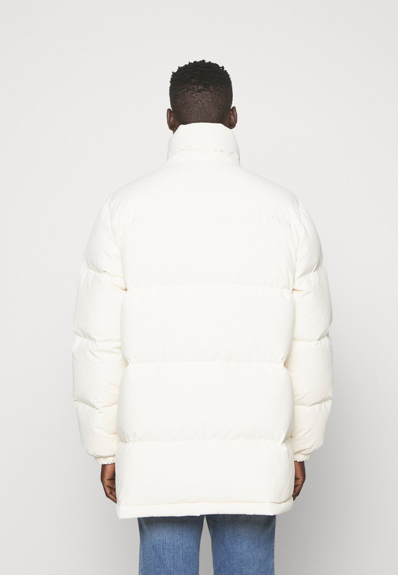 Homme portant un long manteau en duvet blanc et un jean bleu, se tenant de dos contre un fond gris clair uni.