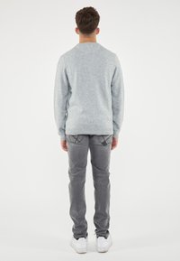Maglione lavorato a maglia grigio chiaro con scollatura rotonda. Abbinato a jeans grigi con dettagli di cucitura delle tasche e sneakers bianche. Sfondo semplice.