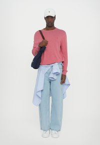 Raudonas megztinis, šviesiai mėlynos plačios kelnės, dryžuotas marškinėliai surišti ties juosmeniu, tamsiai mėlyna per petį nešama rankinė, balti sportbačiai ir balta kepurė.