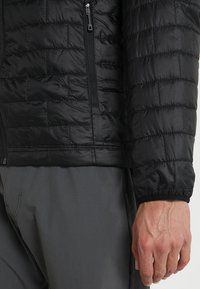 Svart pufferjacka med quiltad design, dragkedjefickor och elastiska ärmmuddar. Lätt material med en slät textur.