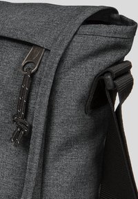 Borsa a tracolla in tessuto grigio con finitura testurizzata, dotata di un lembo pieghevole, una zip marrone e una tracolla intrecciata.