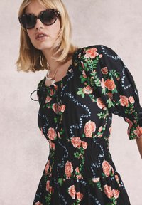 Rochie neagră cu flori, dotată cu un bust smockat, mâneci bufante și modele cu trandafiri. Accesorizată cu ochelari de soare tip cat-eye cu accente florale.