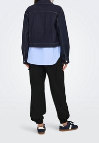 Personne portant une veste en jean foncée, une chemise bleu clair, un pantalon cargo noir avec des poignets élastiques et des baskets bleu marine, debout de dos sur un fond blanc.
