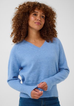 Mujer con cabello rizado que lleva un suéter azul claro con cuello en V y vaqueros, mirando ligeramente hacia arriba con las manos cerca del borde del suéter.