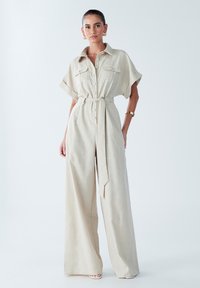 Beige jumpsuit yrityksestä, jossa on lyhyet hihat, napitettu etuosa, kaksi rintataskua ja yhteensopiva vyö vyötäröllä; leveät lahkeet.