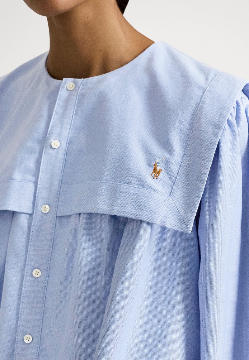 Vrouw draagt een lichtblauwe jurk met knopen, brede kraag en een klein geborduurd polo-spelerlogo op de borst.