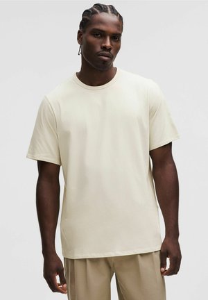 Hellbeige Kurzarm-T-Shirt aus weichem Stoff mit Rundhalsausschnitt, präsentiert an einem Modell, kombiniert mit beigen Hosen.