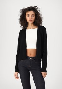Zwarte cropped cardigan met open voorkant, geribde manchetten en zoom. Gecombineerd met donkere jeans en een witte cropped top. Geurkaarsgebreid stoffen.