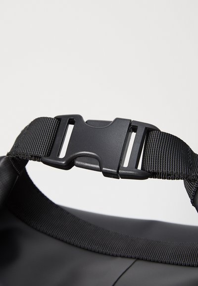 Mustast kott, millel on võrkkonstruktsiooniga rihm, plastist klamber ja reguleeritav riistvara. Sile tekstuur ja matt viimistlus.