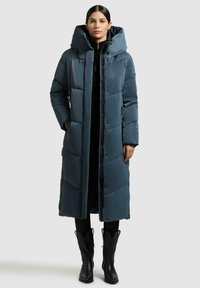 Lang dunvest i mørk teal, med stor hætte, quiltet mønster, lynlås foran og sidelommer; båret over et sort outfit.