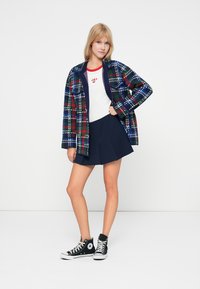 Καρό μπουφάν σε μπλε, πράσινο και κόκκινο πάνω από ένα λευκό μπλουζάκι, συνδυασμένο με ναυτικό φούστα και μαύρα αθλητικά παπούτσια ψηλά.