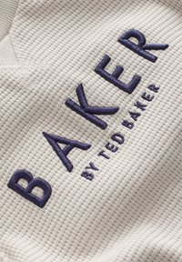 Vit texturerad tyg med ett våffelmönster, med mörkblå broderad text: "BAKER" och "by TED BAKER" under.