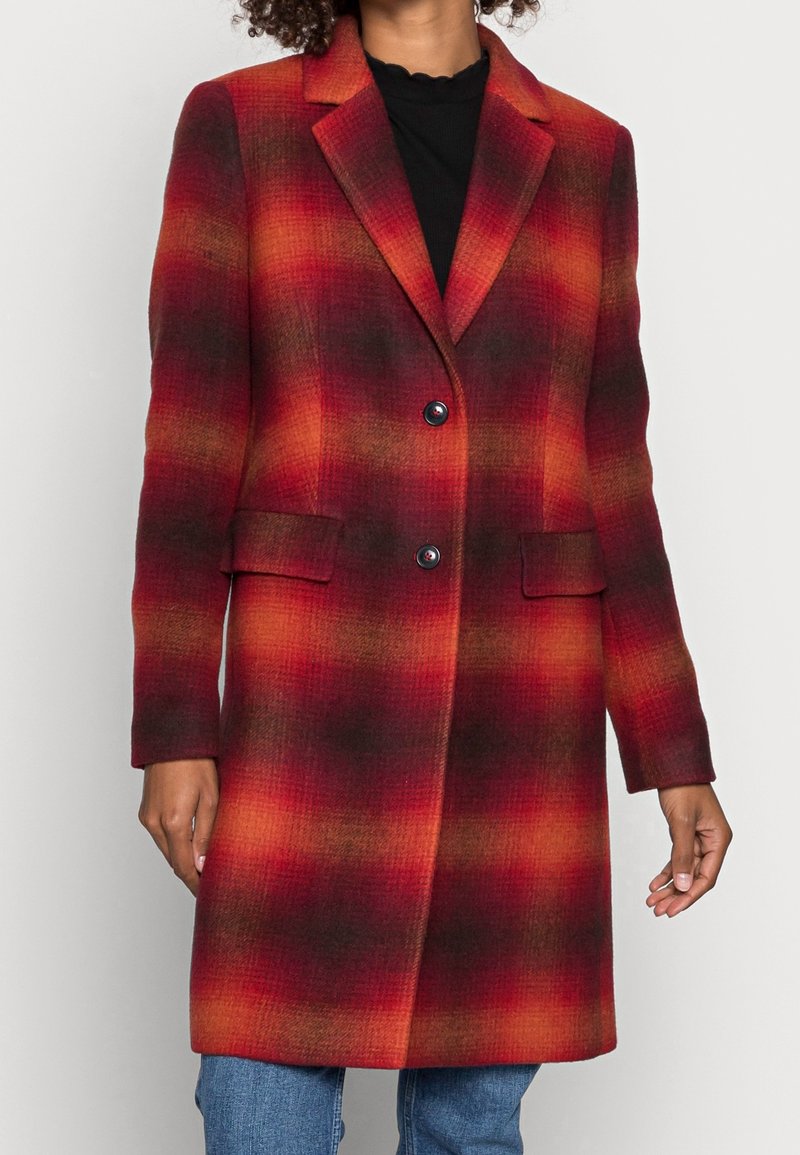 Femme portant un manteau écossais mi-long à dégradé rouge et orange avec deux boutons, des poches à rabat et un col cranté, par-dessus un haut noir.