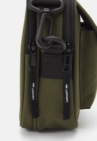 Olivengrønt stofpose med sorte lynlåse, tekstureret overflade og en karabinerspænde. Udstyret med dobbelte lynlåstrækkere mærket "Carhartt WIP."