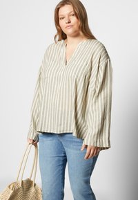 Blusa de linho riscada com decote em V, mangas descaídas e mangas longas. Bege claro com riscas em verde-oliva. Combinada com jeans azul claro.