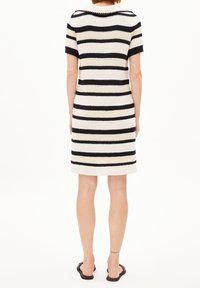 Femme portant une robe courte à manches courtes arrivant aux genoux avec des rayures horizontales noires, beige et blanches, debout pieds nus sur un fond blanc uni.