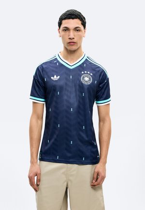 Νεαρός άνδρας που φοράει μπλε ναυτικό ποδοσφαιρική φανέλα Adidas με ανοιχτό μπλε λεπτομέρειες και έμβλημα ομάδας, στέκεται μπροστά σε απλό λευκό φόντο.