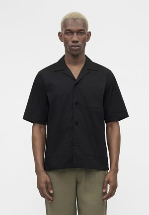 Mężczyzna z krótkimi blond włosami, ubrany w czarną koszulę z krótkimi rękawami zapinaną na guziki oraz oliwkowe spodnie, stojący na jednolitym tle.