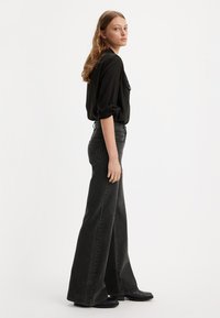 Žena v čiernej košeli na gombíky s vyhrnutými rukávmi, v čiernych vysokých nohaviciach s širokými nohavicami a čiernych topánkach, stojí proti bielej stene.