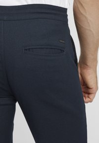 Navy blue pants with a subtle textured pattern, featuring a back pocket and a small logo label near the waistband.