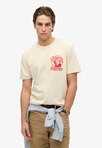Beige bomuld T-shirt med rødt grafisk logo på brystet, parret med khaki bukser og en lyseblå stribet skjorte bundet omkring taljen.