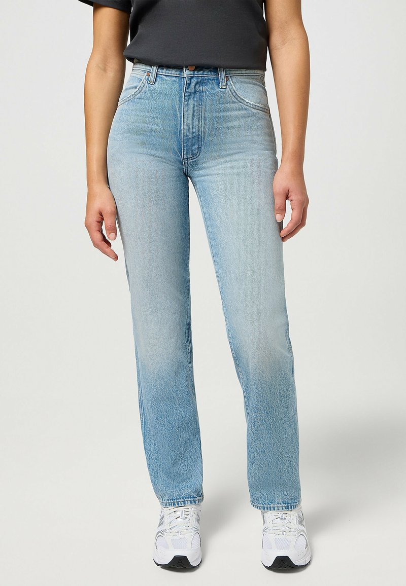 Personne portant un jean taille haute bleu clair, un top noir et des baskets blanches, debout devant un fond uni.
