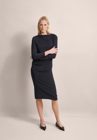 Schwarzes, langärmeliges Kleid mit figurbetonter Silhouette und rundem Ausschnitt. Glattes, dehnbares Material; knielanger Saum. Getragen mit schwarzen spitzen Absatzschuhen.