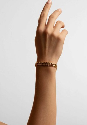 Goldkettchen-Armband mit ineinandergreifenden Gliedern und polierter Oberfläche. Getragen an einer Hand mit neutraler Nagelfarbe vor einem hellen Hintergrund.