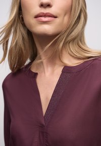 Vinrød bluse med V-hals og tekstureret kant, flydende stof og en glat finish. Nærbillede fremhæver halsudskæringen og materialet.