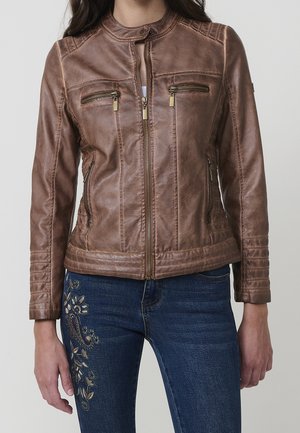 Veste en cuir marron avec fermeture éclair à l'avant, poches zippées sur la poitrine et les côtés, portée sur un jean bleu avec une broderie florale sur la cuisse gauche.