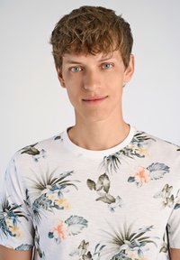 Valkoinen lyhythihainen t-paita, jossa on trooppinen kukkakuosi vihreillä lehdillä ja oransseilla kukilla, valmistettu pehmeästä ja kevyestä kankaasta.