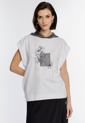 Kobieta ubrana w biały, bezrękawnik z czarnym kwiatowym i geometrycznym wzorem oraz czarną spódnicę, stojąca na jednolitym białym tle.