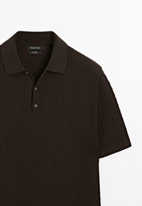 Kortærmet polo T-shirt i mørkebrun, lavet af bomuld med ribbet tekstur. Har en krave og to knapper ved halsudskæringen.