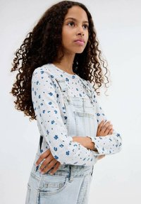 Long-sleeve white top with blue floral print, paired with light blue denim overalls featuring adjustable straps and front pocket.