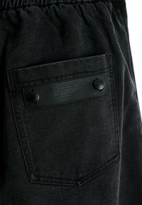 Pantalones de mezclilla negros con una cinturilla elástica, un bolsillo trasero con una etiqueta de cuero negro texturizado y acentos de botones redondos.