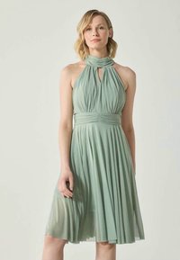 Pea rohelises halter-kaelusega kleit, millel on voldid vööst hemline'ni ja sobiv bodice koos ronnendatud talje detailiga.