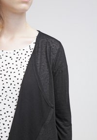 Cardigan nero in materiale leggero, con apertura frontale, tessuto strutturato e vestibilità rilassata, abbinato a una maglietta bianca con triangoli neri.