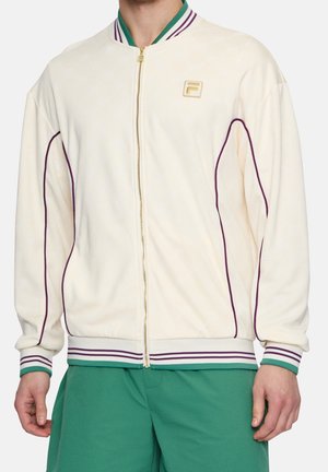 Mand iført en cremefarvet lynlåstrøje med lilla piping og stribede manchetter i grøn og lilla, kombineret med teksturerede grønne bukser.