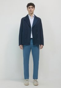 Giovane uomo in piedi, che indossa un blazer blu navy a doppiopetto, una camicia a righe azzurro chiaro, pantaloni verde acqua e scarpe bianche stringate, davanti a uno sfondo semplice.