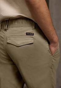 Homme portant un pantalon beige décontracté avec une ceinture élastique et une rabat de poche, main dans la poche latérale, montrant l'étiquette "Street One Men".