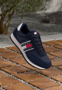 Scarpe da ginnastica blu navy con tessuto testurizzato, accenti bianchi e strisce rosse. Presentano un colletto imbottito e chiusura con lacci, esposte su piastrelle di mattoni.
