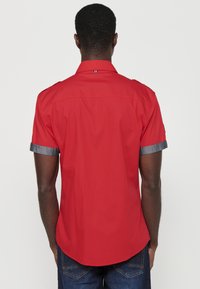 Camisa corta de manga roja con cuello abotonado, que presenta acentos de denim en los puños enrollados y un dobladillo curvado.