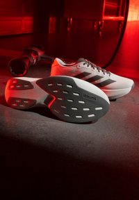 Paar weiße Laufschuhe mit schwarzen Streifen und Continental-Gummisohlen auf Betonboden, mit roter Beleuchtung und Trainingsgeräten im Hintergrund.