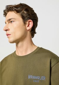 Oliwkowa bawełniana koszulka z okrągłym dekoltem i niebieskim logotypem "WRANGLER" na lewej piersi, o gładkiej strukturze.